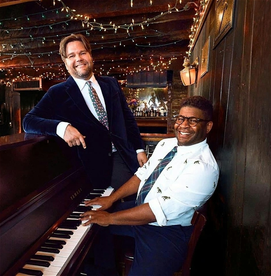 Image is of two people at an upright piano in a bar with fairly lights strung across the ceiling, both wearing suits and ties and smiling to the camera. One person is seated at the piano with their hands on the keys the other is stood slightly behind, leaning on the end of the piano.