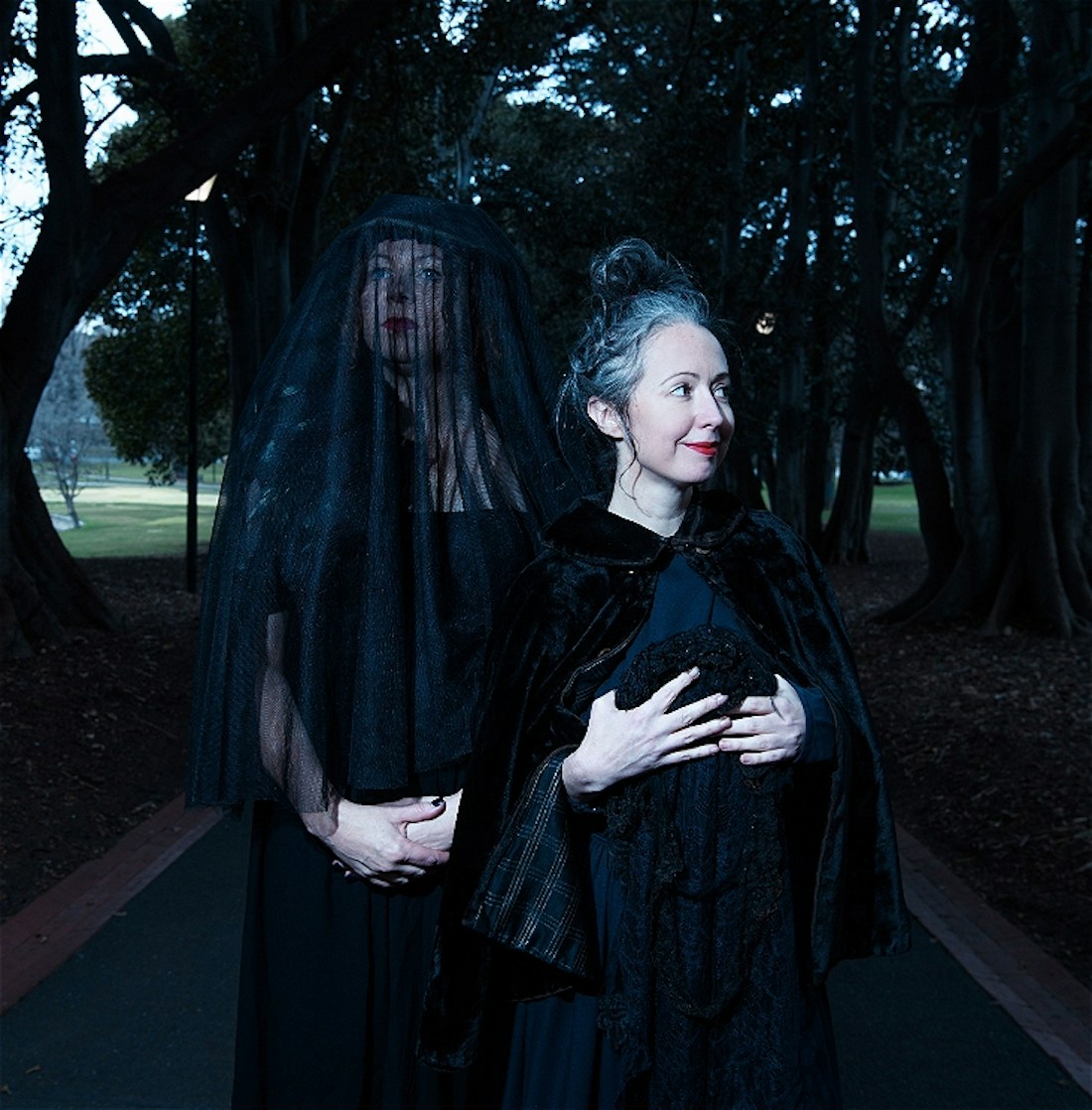 Two women in funereal costume pose. One is wearing a veil. The other looks off into the distance.