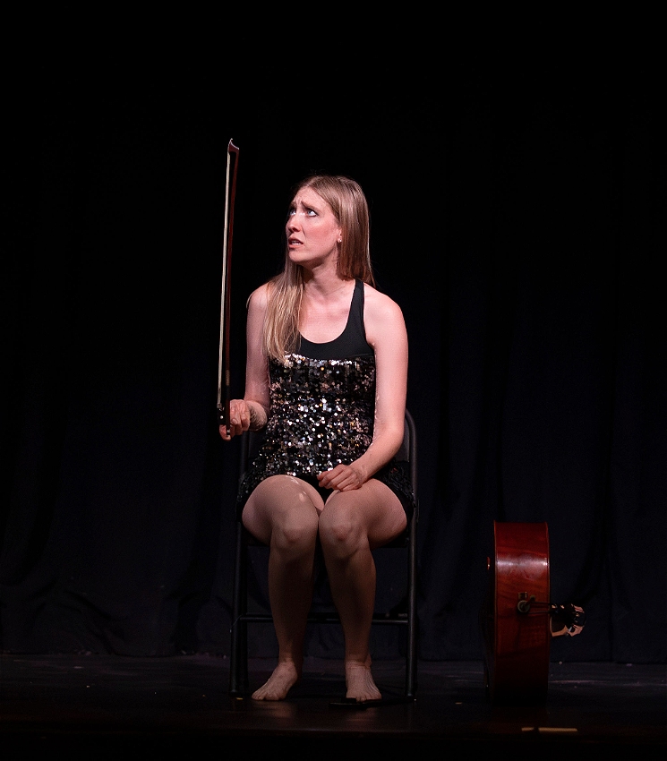 A performer with long, blonde hair sits on a chair on a black stage, dressed in a sparkly black mini dress and delicately balancing her cello bow on a single index finger. Her face looks up to the bow, looking very unsure about what's happening.