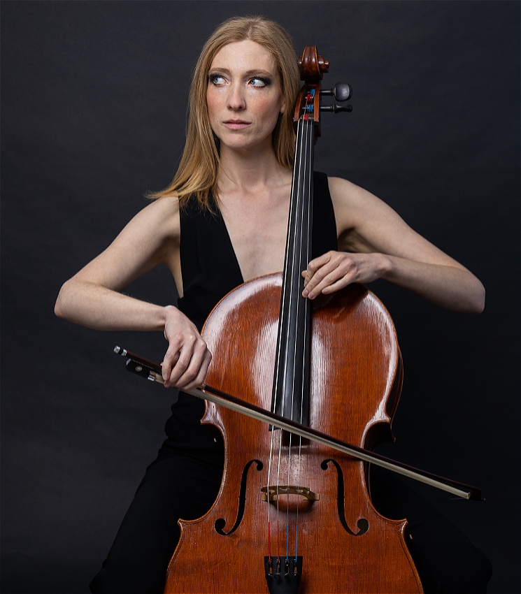 A cellist with long blonde hair and formal black attire sits playing her cello. Her eyes looking sideways as if on the lookout for something.