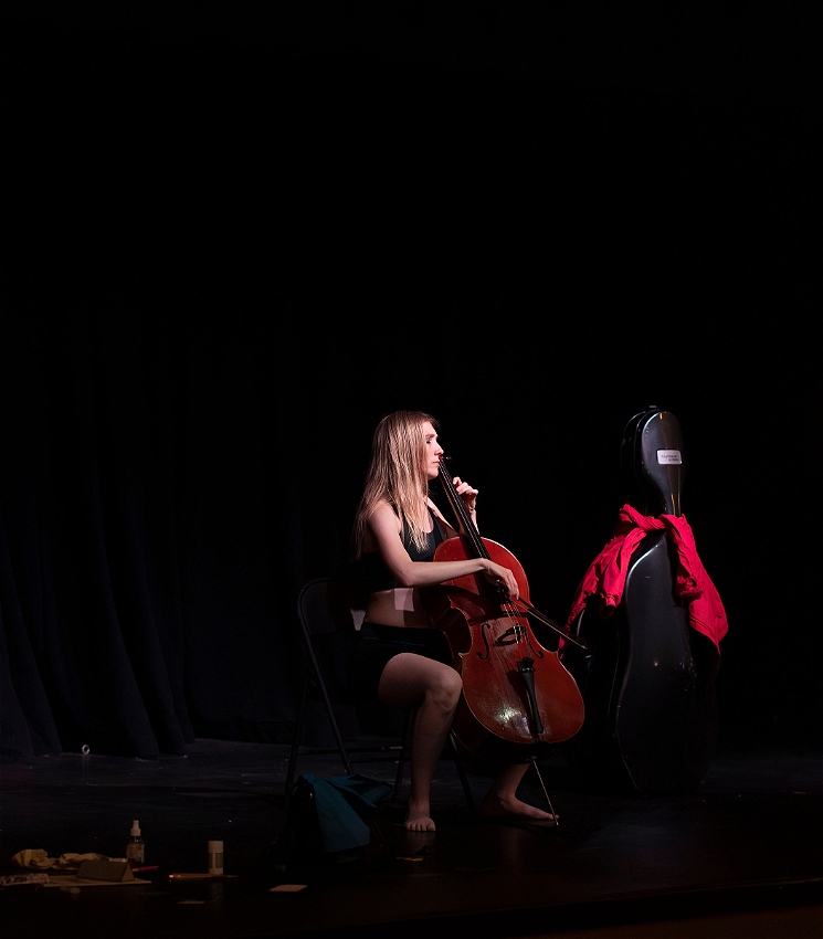 A performer with long, blonde hair sits on a chair on a black stage, lit by a spotlight and dressed in a black top and shorts. She peacefully plays a cello amid a clutter of small items and a cello case on the stage - a calm after a storm.