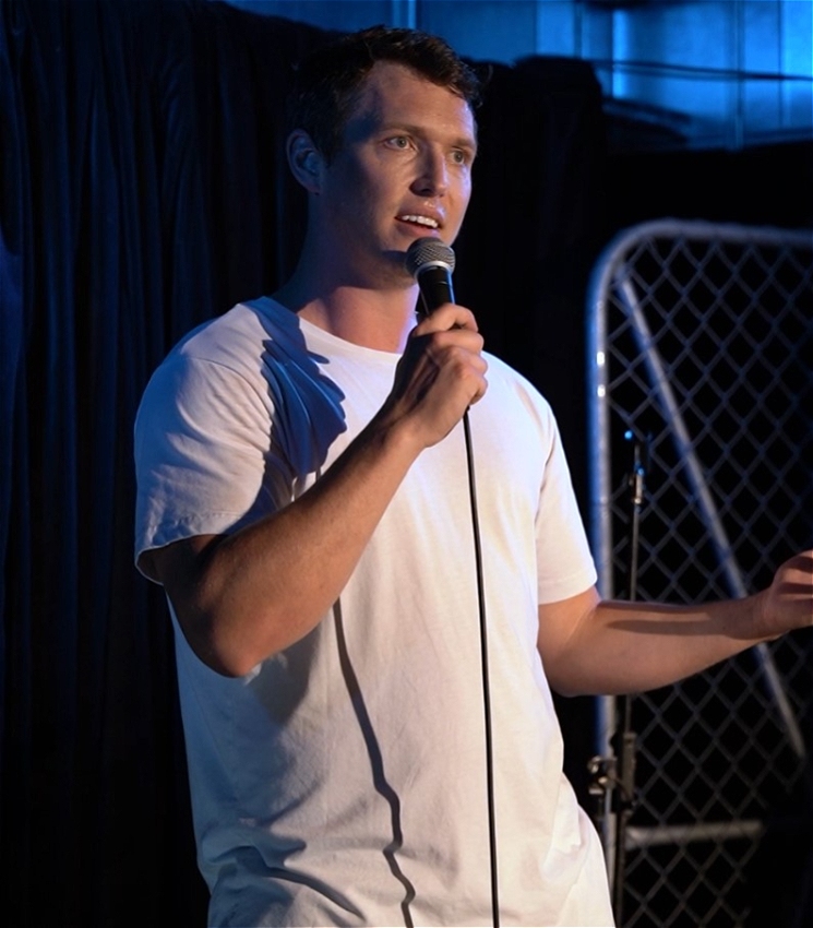 A man wearing a white tshirt stands on a stage talking into a microphone. There is a black curtain and wire fence behind him.