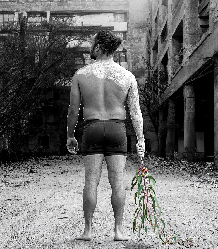 A black and white photo of an acrobat with their back to the camera. The skin on the top of their back and right arm are covered in white ochre, they are holding a branch of eucalyptus leaves by their side which are green and pink in colour.