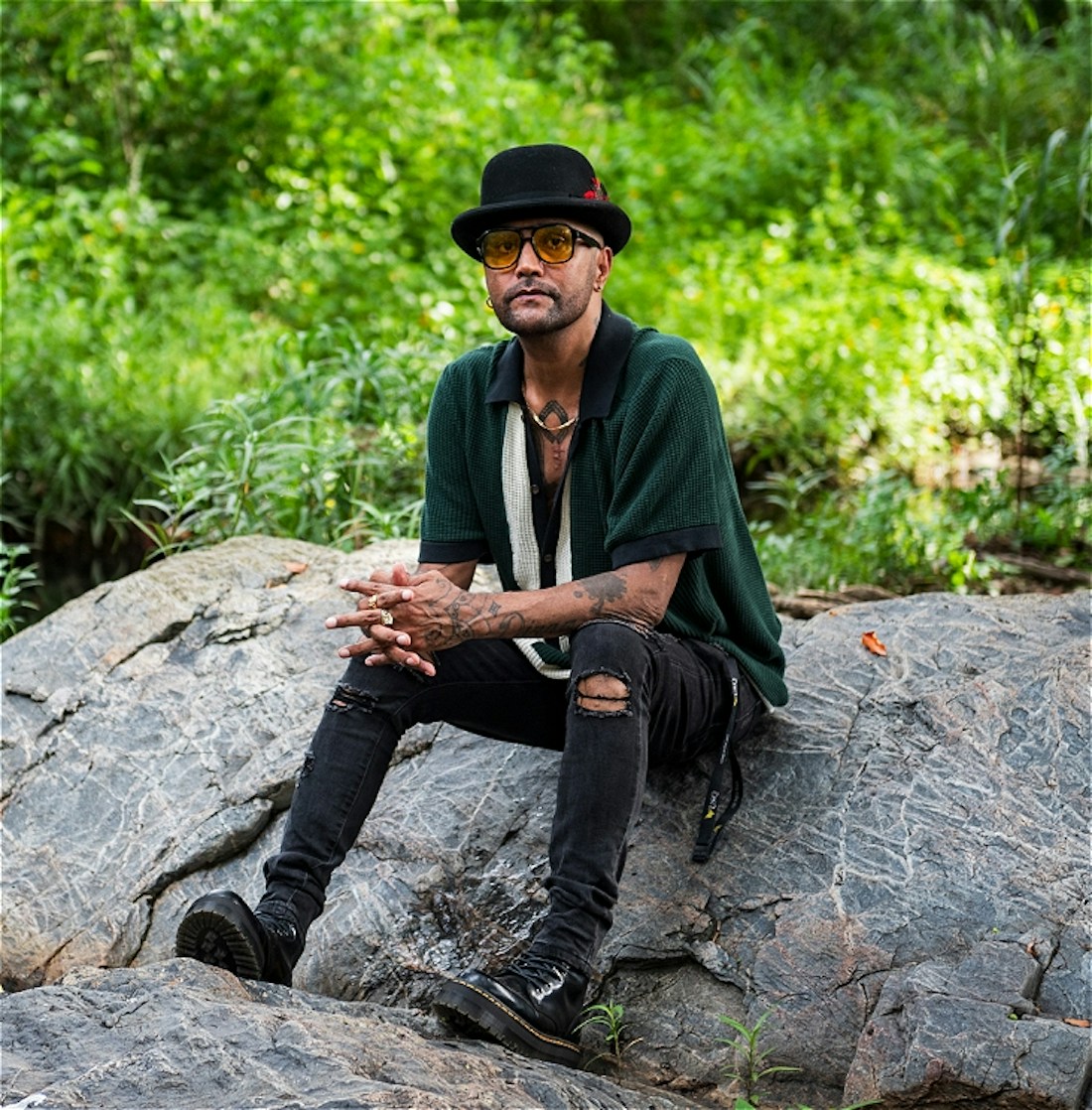 JUNGAJI, A Gugu Yalanji first nations artist, wearing a black fedora hat, green shirt with torn black jeans. He's sitting on a rock by a river stream amongst the beautiful green forest.