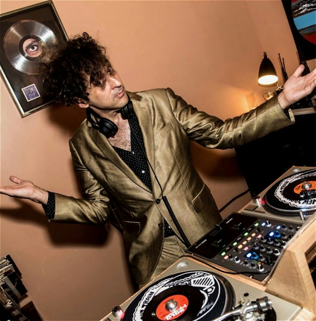 A man wearing a gold suit jacket stands in front of some DJ decks. He is in a room with light orange walls and wall and vinyl records as decoration on the walls. The photo is tilted and the man stares at the camera with a playful expression.