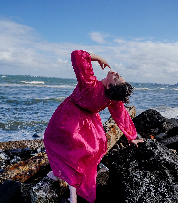 Ilana Charnelle, a white woman with short brown hair and bright red lipstick, is pictured on a rocky outcrop with the ocean visible in the distance. She leans back at a dramatic angle against the rock, with one hand on the rocks and the other striking a pose above her head.