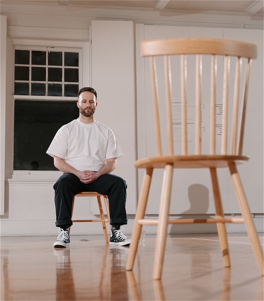 Man with a white t-shirt, navy pants, and Chuck Taylor shoes sits on a wooden chair. Opposite him is an empty chair, and behind him is a darkened window. This is in a gallery with wooden floors and white walls.