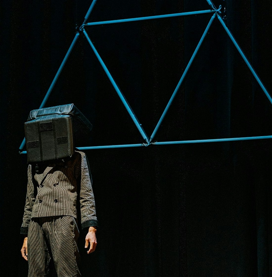 A person in a suit stands in front of a neon blue triangle with a big black box on their head