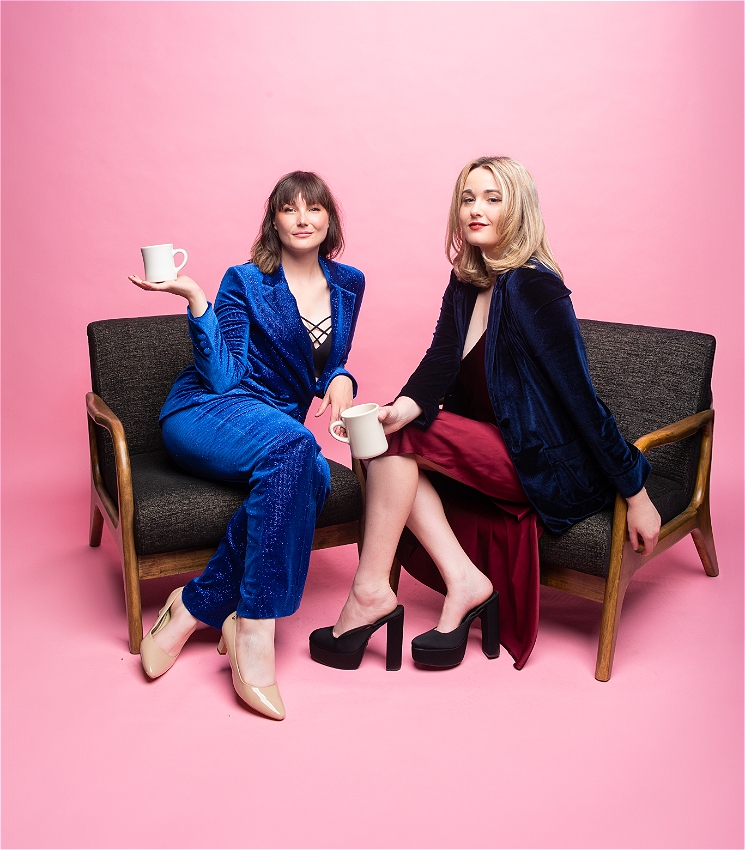 Two women sit in armchairs against a bubblegum pink background. One has brown hair and is in a blue suit the other has blonde hair and is in a dress and blazer. They both hold white coffee mugs and looking at the camera with a confident look on their faces.