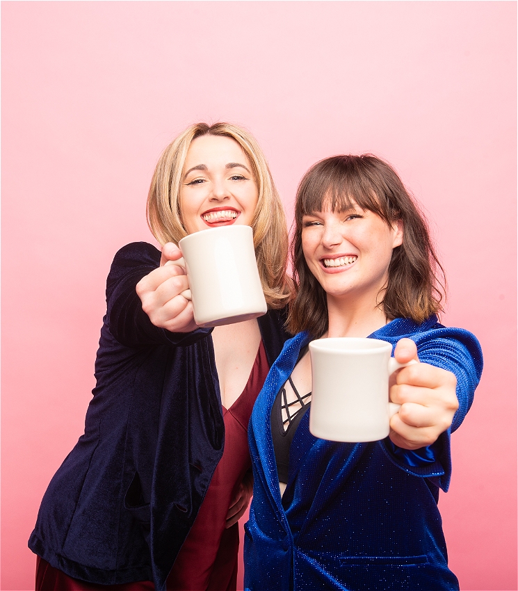 A blonde and brunette woman dressed in suits hold white mugs towards the camera while smiling cheekily against a baby pink background.