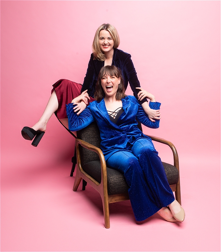 A brunette woman in a blue sparkley suit sits in a chair while open mouth laughing. A blonde woman is sitting on a stool behind her in a dress and blazer with a wide smiles. The two are in front of a baby pink background.