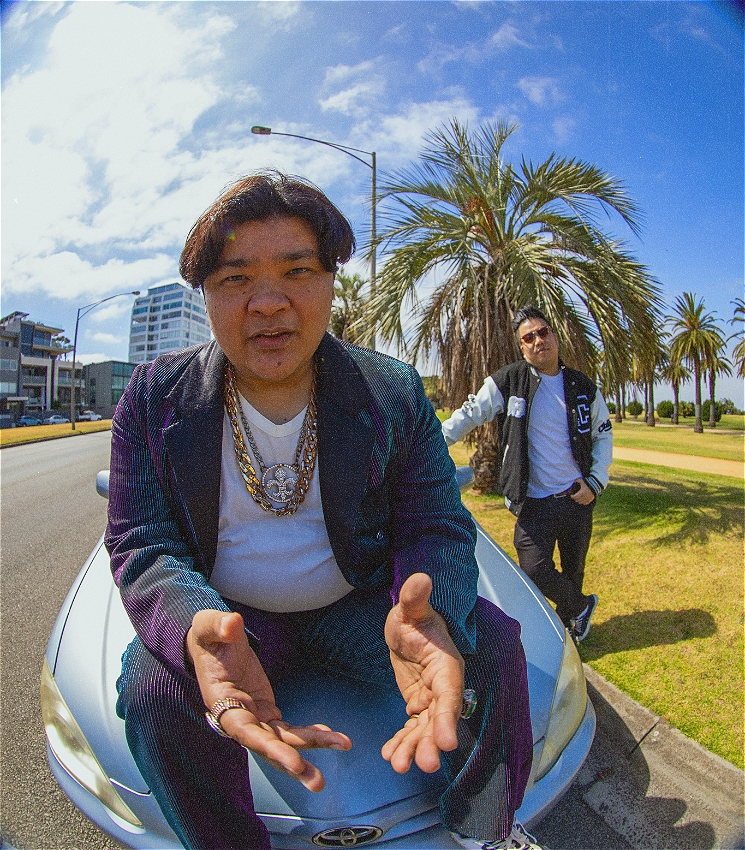 A man in a blue and purple corduroy suit with large chains around his neck sits on a 2006 Toyota Corolla taken with a fish eye lens. Another man is behind him leaning on the side of the car in a varsity jacket. The car is parked along a street lined with grass and palm trees.