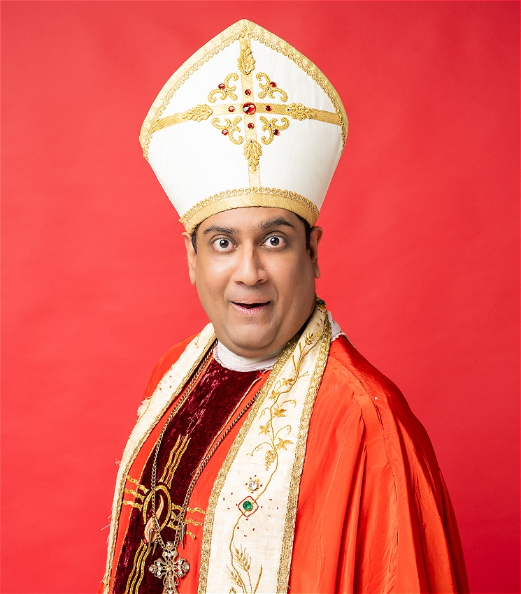 Sri Lankan Man in red and white pope costume adorned with an ornate cross necklace, cheekily smiles at camera in front of bright red background.