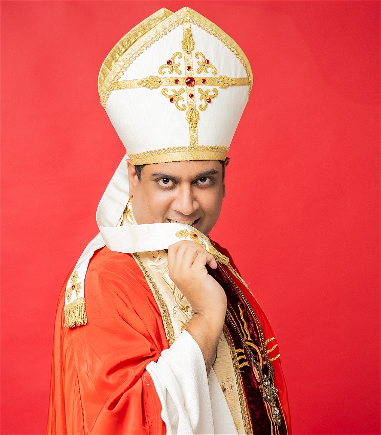 Sri Lankan Man in red and white pope costume adorned with an ornate cross necklace in front of bright red background. He sexily bites at a tassel from his hat while sultrily looking into the camera.