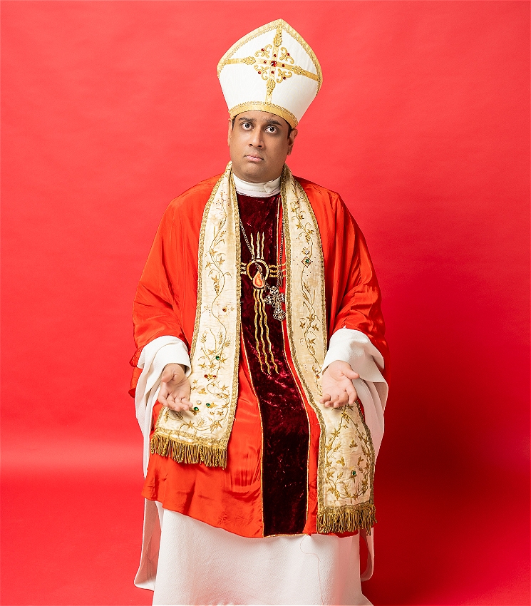 Sri Lankan Man in red and white pope costume adorned with an ornate cross necklace, sits forlornly in front of bright red background.