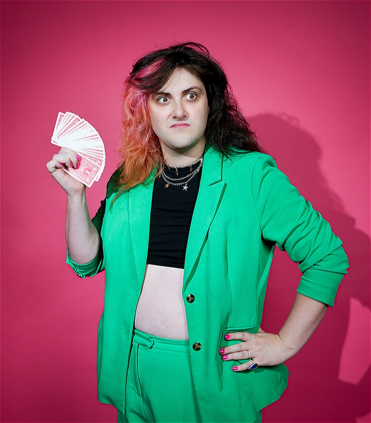 Performer, Tricksy, wearing a bright green blazer and pants, black crop top and holding a spread a playing cards in her right hand in front of a bright pink backdrop. She's looking off to the right with a bit of a stank face.