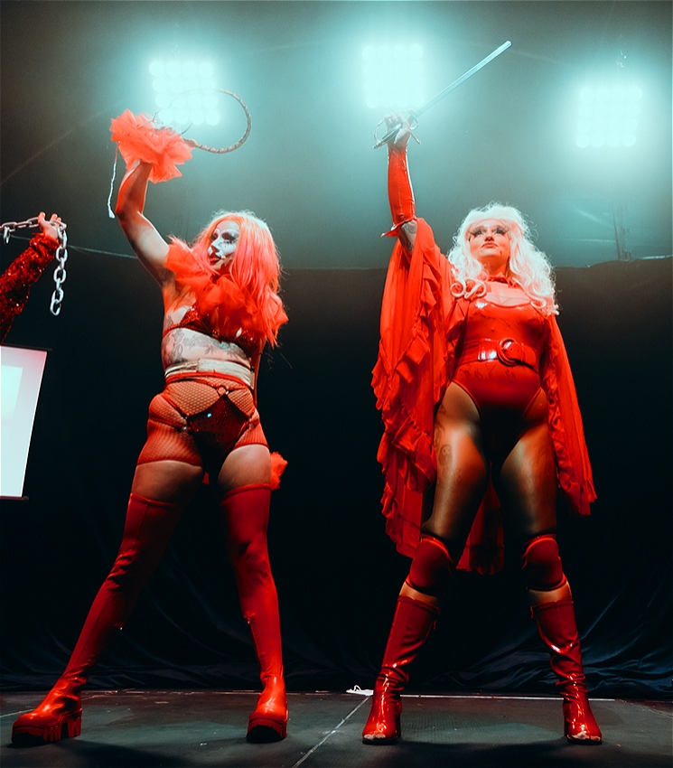 Two performers in red bikinis and thigh high boots stand on stage, holding a whip and a sword above their heads triumphantly.