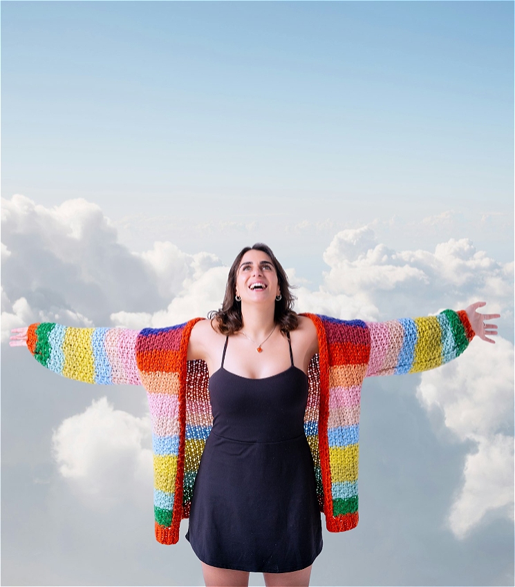 Ashley (woman with olive/Caucasian skin, brown, wavy shoulder length hair, hazel eyes, wearing small silver hoop earrings) stands in front of a background of white clouds with her arms outstretched to show her wingspan (she is tall and has long, gangly arms). Her head looks up towards the sky behind/above her and she has a big, open mouthed smile as her eyes look up. She wears a black, spaghetti strap dress that comes to her thighs, and a large, woollen, knitted cardigan that has horizontal multi-coloured, thick stripes knitted onto it (forest green, a burnt orange, a lime green, baby blue, baby pink, a light mauve and a deep indigo).