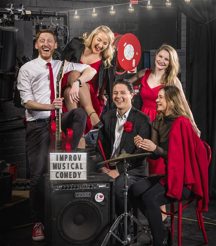 A group of five performers wearing red, black and white are sitting surrounded by musical instruments in a black box theatre space, laughing together.