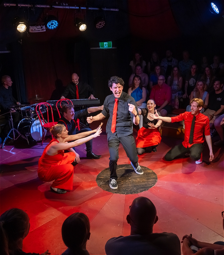 A man is singing passionately on stage surrounded by four performers in a tent-style round theatre.