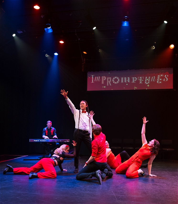 A man is posing surrounded by four performers with their hands in the air and a musician playing piano. A sign on the curtain behind the performers reads 