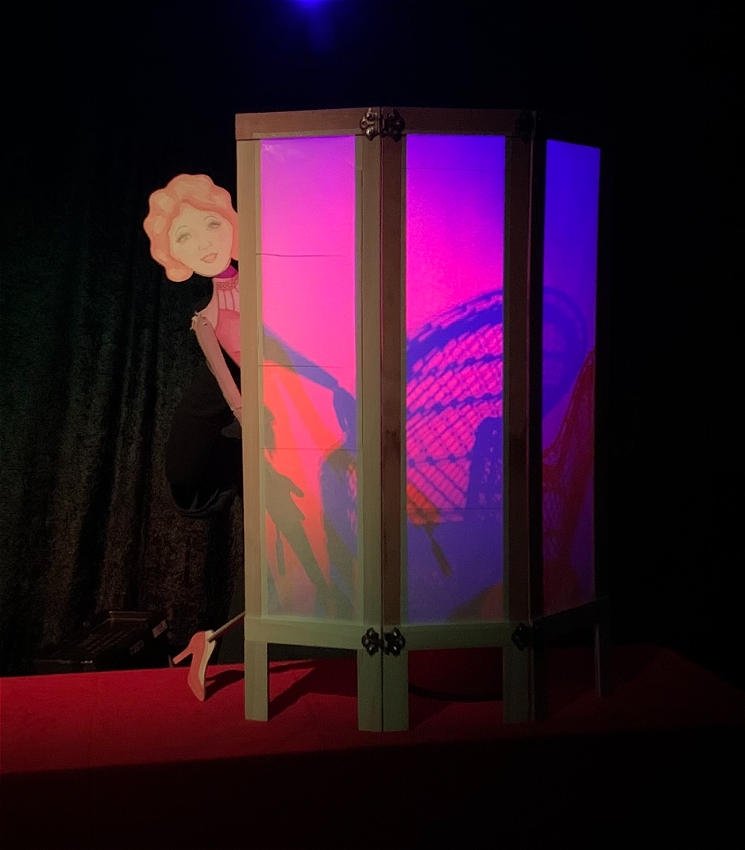 A three-panel room-divider screen is backlit with pink and purple light. A shadow of a cane chair is cast on the screen. A painted cardboard woman with red wavy hair leans out from the side of the screen and reveals her red stiletto at the bottom.