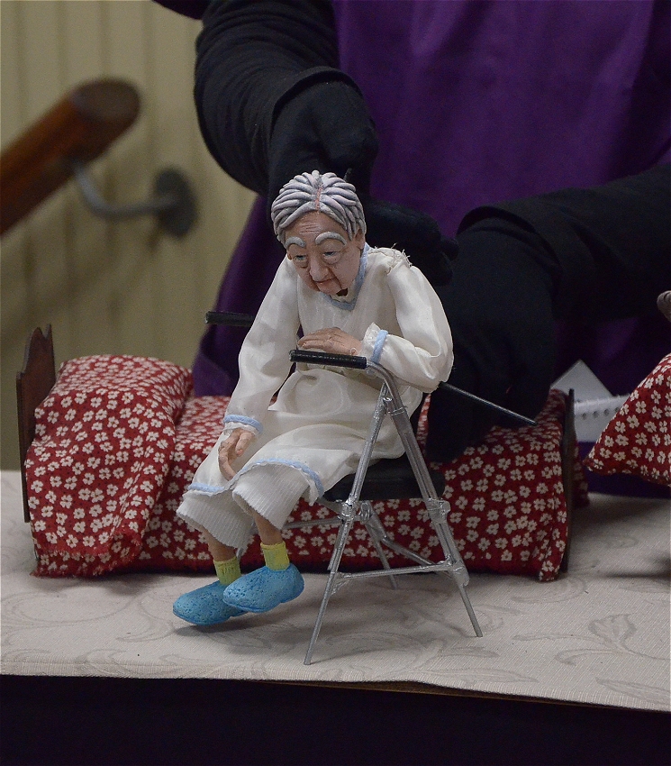 A small old lady puppet is looking down as she sits on a silver walking frame. She is wearing a white nightie and blue slippers with short yellow socks. Behind her is a bed covered with a red and white-flowers bedspread