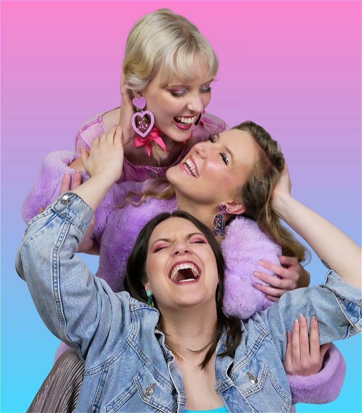 Three white women laugh widely with their arms wrapped around each other and smiling at one another. Their heads are stacked on top of one another. The woman at the top, Rosie, has platinum blonde hair and is wearing her hair in plaits with bright pink ribbons and heart-shaped earrings. The woman in the centre, Mim, has dark blonde hair, and is wearing sparkly earrings and a fluffy purple coat. The woman at the bottom, Jemma, is wearing a blue denim jacket and maroon eyeshadow and lipstick. Behind them is a gradient of bright pink, purple and blue, reminiscent of the bisexual flag.