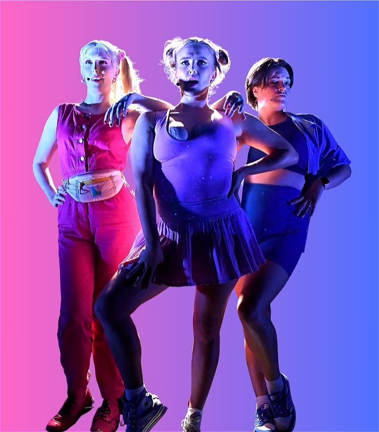 Three white women pose with serious faces, hands on hips and arms leaning on one another's shoulders. The woman on the left is wearing a pink jumpsuit and bum bag, the woman in the centre is wearing a purple bodysuit and tennis skirt, and the woman on the right is wearing blue shorts, crop shirt and oversized button-up.