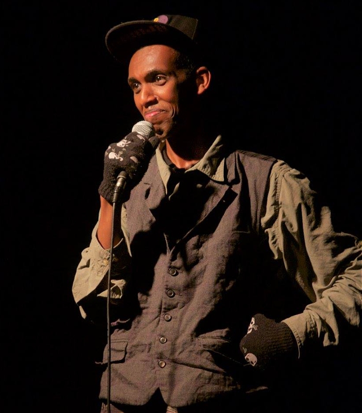 A man is standing on stage, holding a microphone close to his mouth. He is wearing a dark vest, a long-sleeved shirt, a black cap, and gloves with skull patterns. The background is dark, emphasizing the spotlight on him.