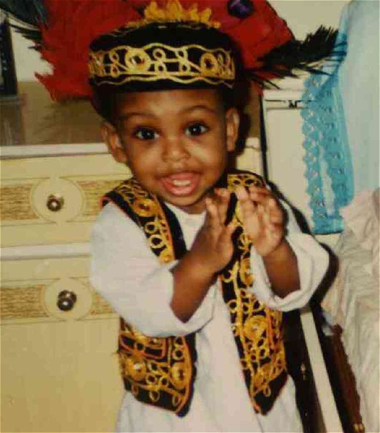 A young child dressed in traditional Somali attire is clapping and smiling at the camera. The child is wearing a white outfit with an intricately designed black and gold vest and matching hat, adorned with colourful feathers. The setting appears to be a cozy bedroom with a bed and a dresser in the background.