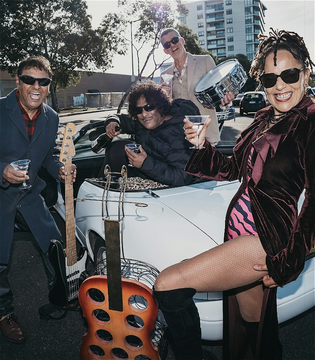One woman and three men, all laughing and drinking cocktails, are gathered in and around a white, convertible car. The woman and two men are people of colour. One man holds a bass guitar. Another, a snare drum. The woman wears a long, burgundy, velvet coat over a pink and black, zebra striped swimsuit with over-the-knee black suede boots.