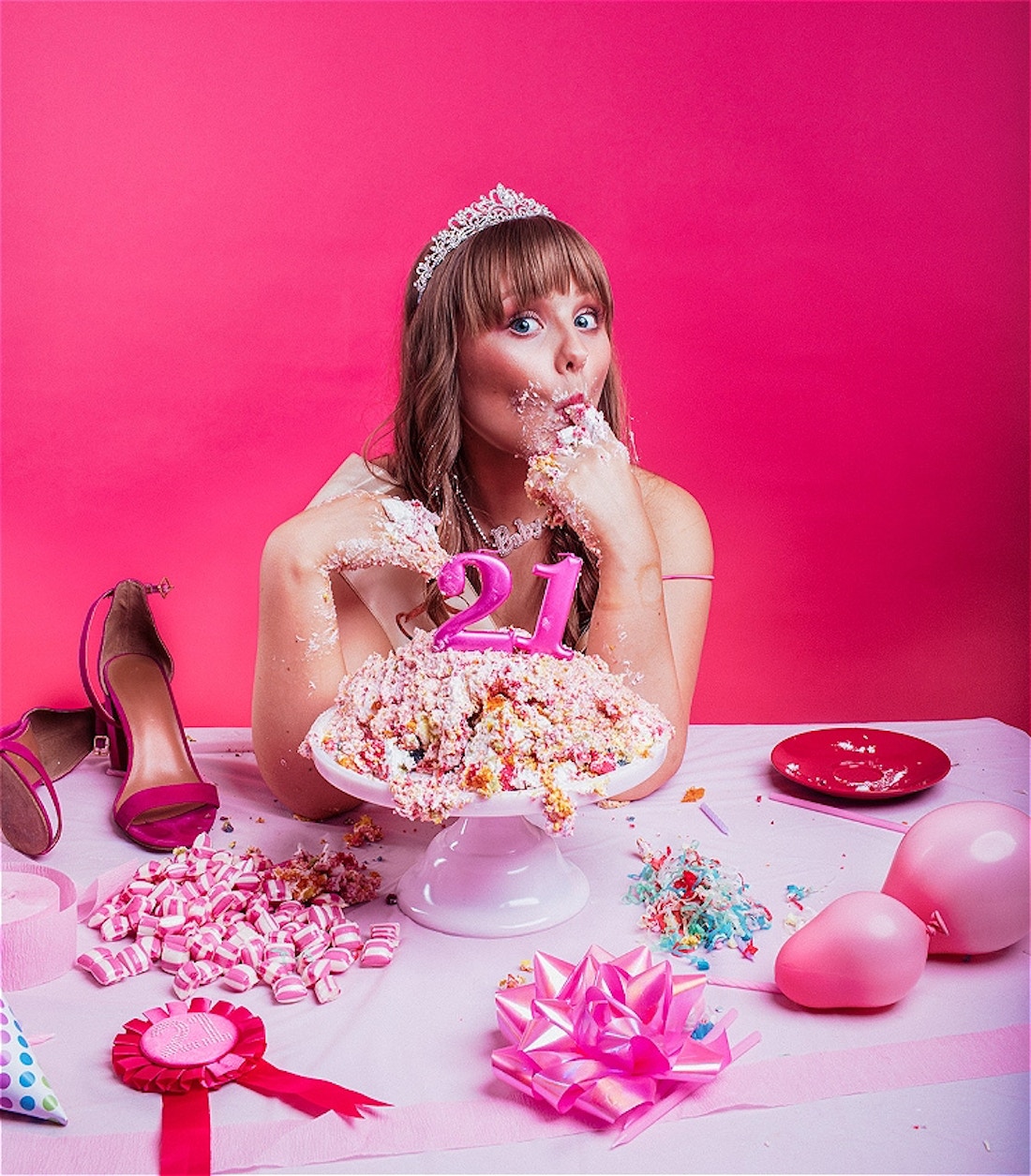 A young woman wearing a tiara is sitting at a messy table eating a birthday cake with her hands. Her face and hands are covered in cake and icing. The table is littered with candy, streamers, balloons and ribbons with candles of the number '21' sitting on top of the destroyed cake.