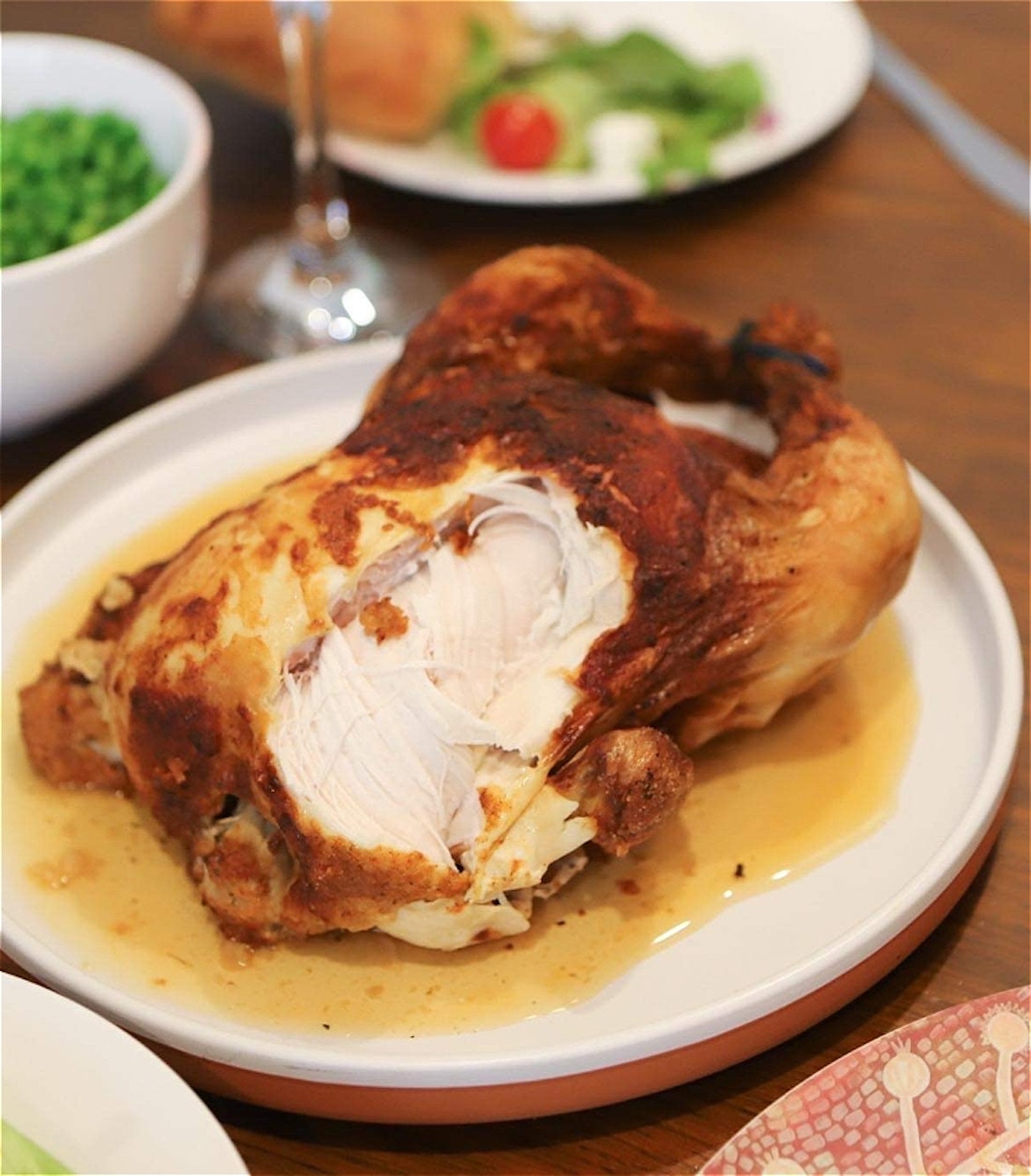 Central to this image is a close up of a rotisserie cooked chicken on a white plate with its orange-ish chicken juice pooling underneath it, all on top of a wooden dining table background. A piece of the chicken breast has been ripped off by an unseen person revealing the sinewy white meat within – it doesn't look very appetising. Placed behind and in front of the chicken, slightly out of focus, are side dishes; there are bright green peas, a green bottle of sparkling water, the stem of a wine glass, and then another plate with some salad greens, a red tomato, and what could be the missing piece of chicken breast – a blurry dinner knife is next to this plate. At the bottom of the image there are the edges of two more plates suggesting the table is set for a normal, suburban, family dinner.