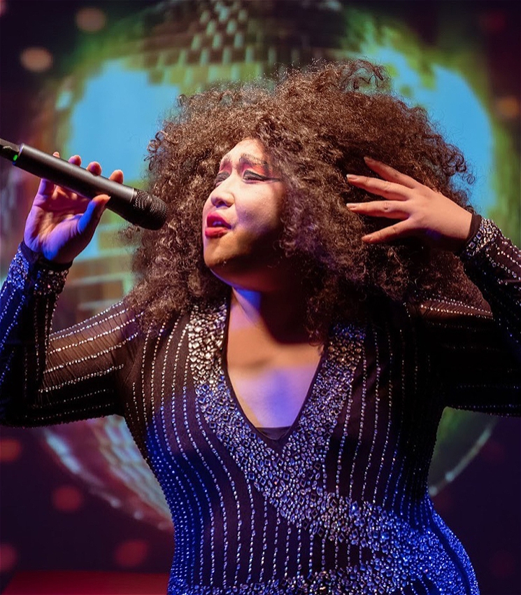 A performer dressed in a tight black outfit covered in silver rhinestones holds a microphone to their face with one hand and holds back their long curly hair with the other in a very dramatic singing pose. They are situated in front of a large projected image of a disco ball.