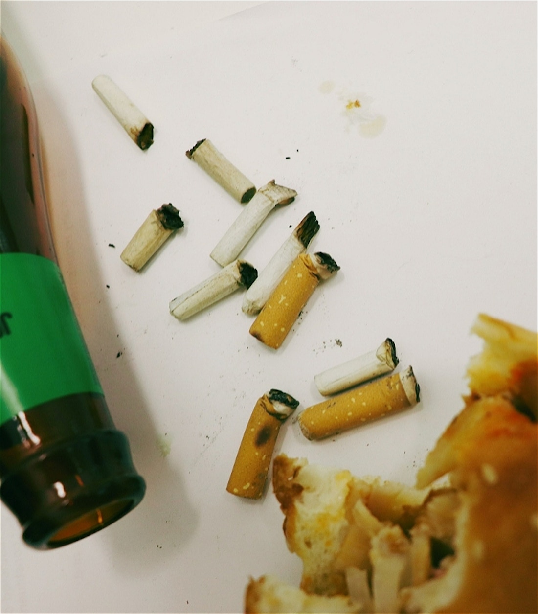 Dirty cigerette butts litter a white background stained with ash. An empty beer bottle with a green label sits on it's side. A kebab with a bite taken out of it is in the corner of the frame.