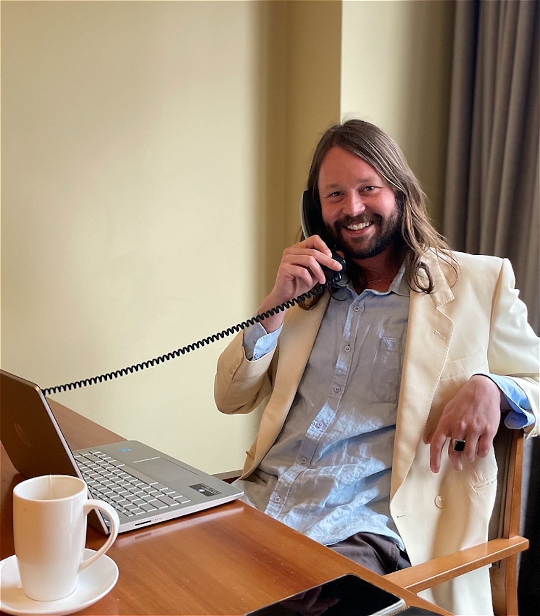 The artist dressed formally sitting at a desk holding a telephone with a laptop and coffee cup in front of him.