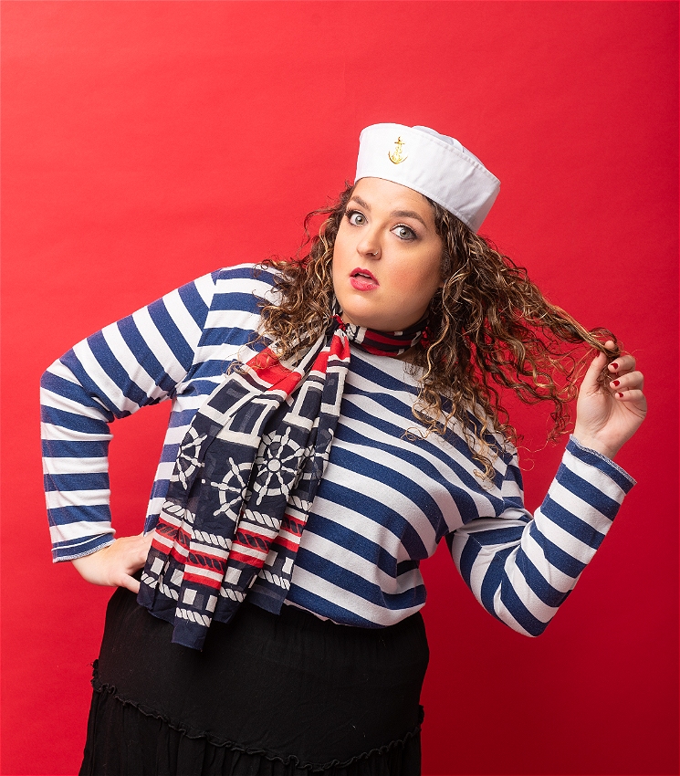 A Caucasian woman in a sailor costume looks to camera and plays with her curly brown hair.