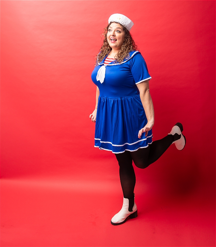 A caucasian woman wearing a blue sailor dress looks off-camera and poses in a vintage way with one leg in the air.