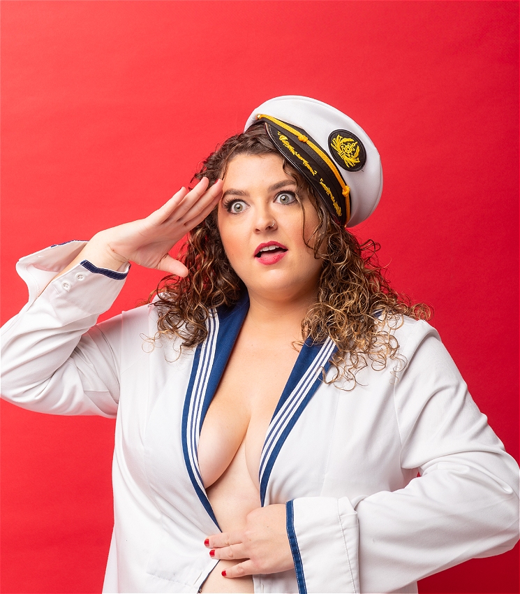 A causcasion woman with curly brown hair salutes and then stares off into the distance with a silly expression on her face. She's wearing a very revealing sailor shirt and a captain's hat.