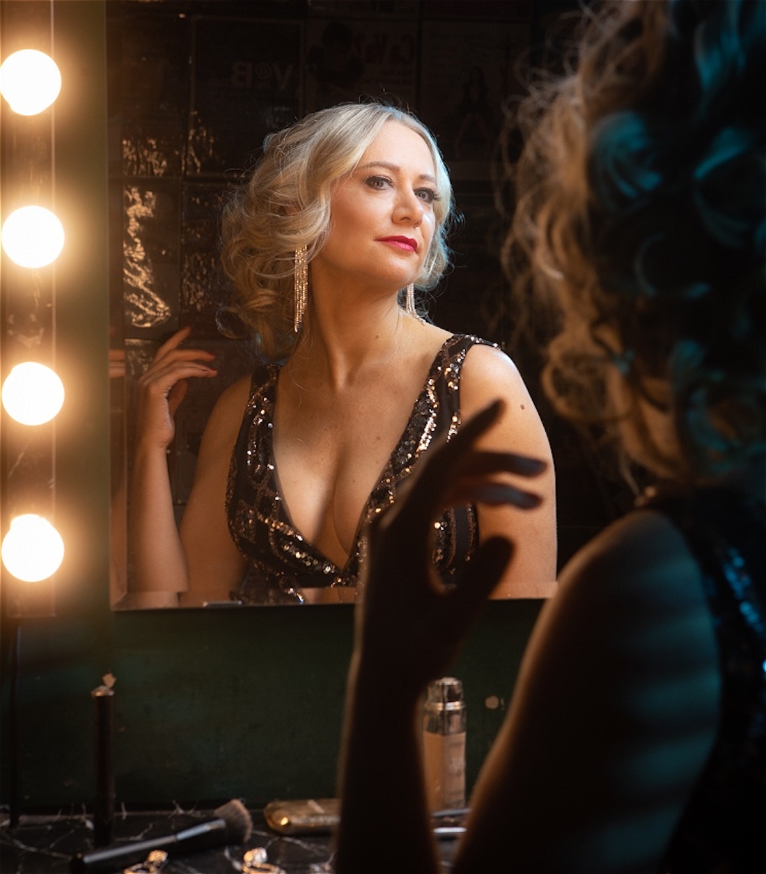Performer Clare Elizabeth Dea, a Caucasian adult woman is sitting in front of a dressing room mirror with diva lights around it. She is wearing a black sequin dress, long silver dangling earrings and her blonde hair is short and curled. We see a dark image of the back of Clare's hair, shoulder and arm and in the mirror see a diva character looking posed but not all together.