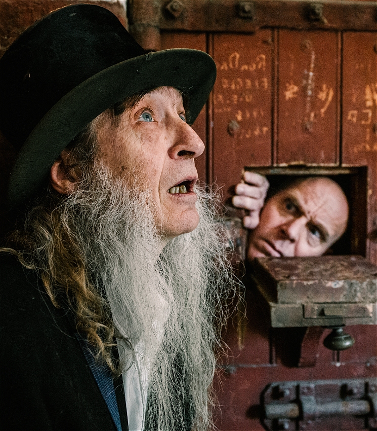 An old disheveled priest-like character with a top hat and long beard, is outside an old prison door. He's looking upwards perhaps in prayer. Through an opening in the prison door, we see the face of second man who seems alarmed.