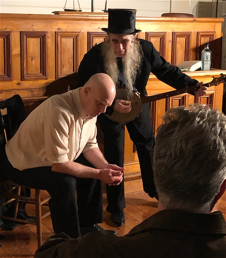 Photographed on stage, a man in a white shirt and black trousers, sits on a chair looking pensive while a banjo playing priest-like character, leans over him in mockery.