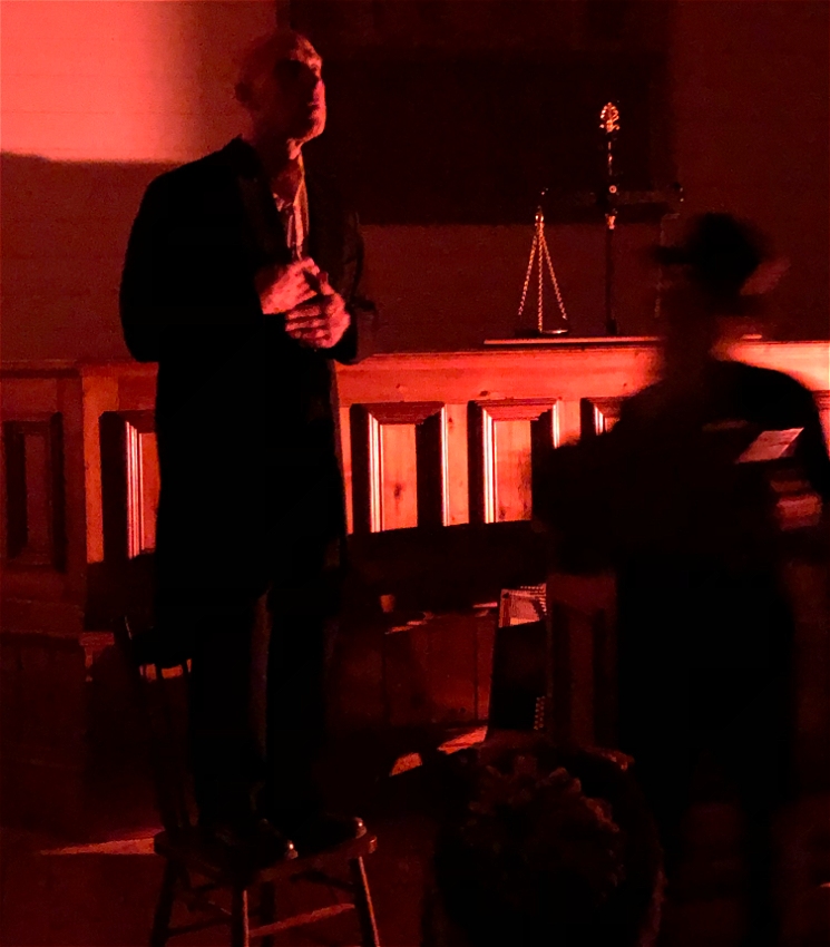 Photographed on stage, a character stands on a chair looking skywards perhaps in prayer. The scene is bathed in bright red light. An out of focus second character, with a banjo, is moving past him.