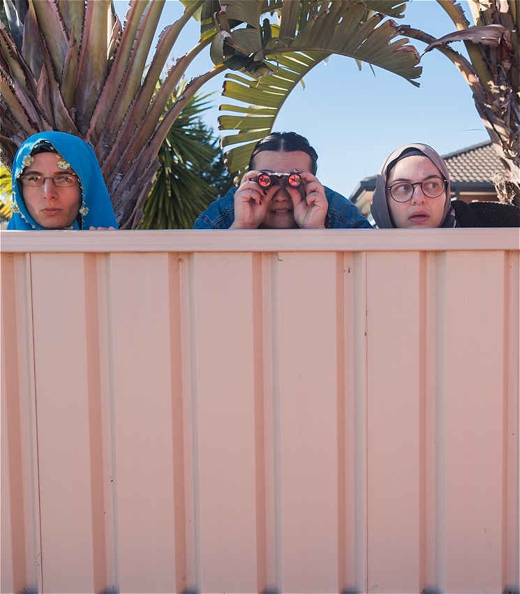 The tops of the faces of three women looking over a suburban metal fence spying on their neighbours. They are dressed in casual attire. The woman in the middle is looking through a pair of binoculars, the woman on the right appears shocked, and the woman on the left looks curious and suspicious.