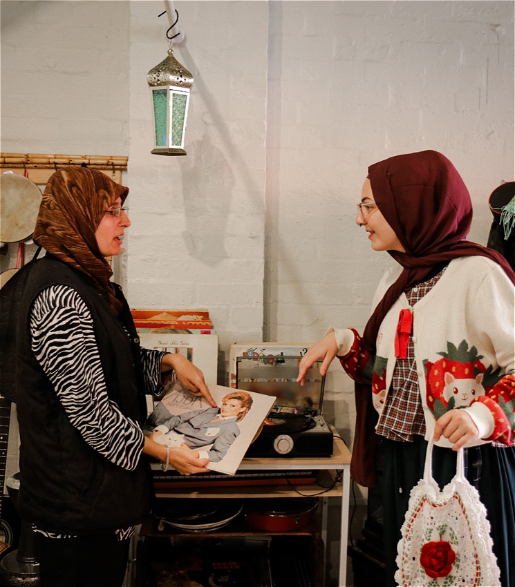 There are two visibly identifiable Muslim women talking in a shop. There is a record player beside them. One of the ladies holds a record and points to it, while the other smiles.