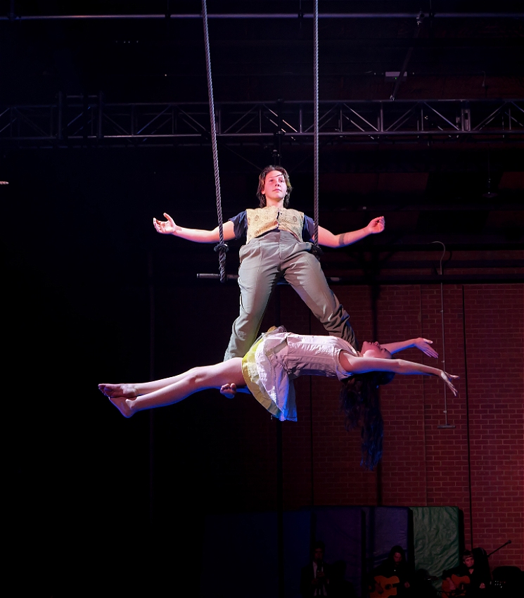 A circus artists sits on a trapeze and leans back with arms wide. A second circus artists lies across their feet with arms out stretched.