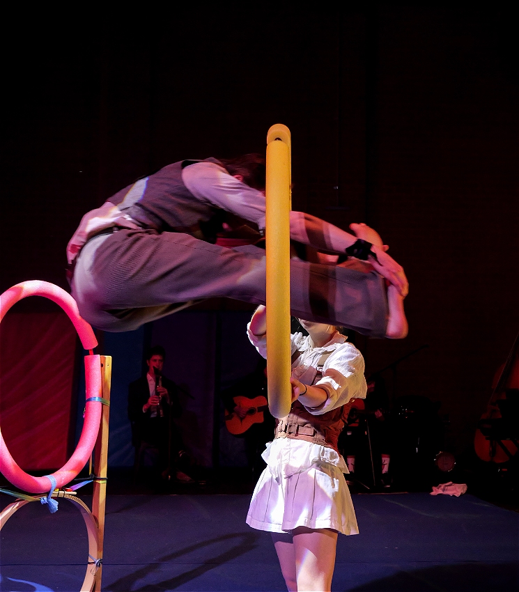 A circus artist holds a yellow hoop at head height. A second circus artist is mid-jump through the middle of the hoop, with hands reaching towards their feet in a folded shape.