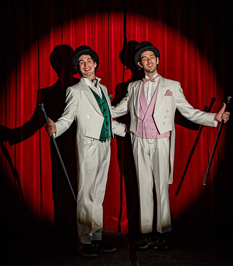 Two brothers stand smiling at the camera with arms wide open, wearing white tailcoat suits with top hats and canes in front of a spotlight-lit red curtain. One brother wears a green waistcoat and bowtie, while the other's is pink.