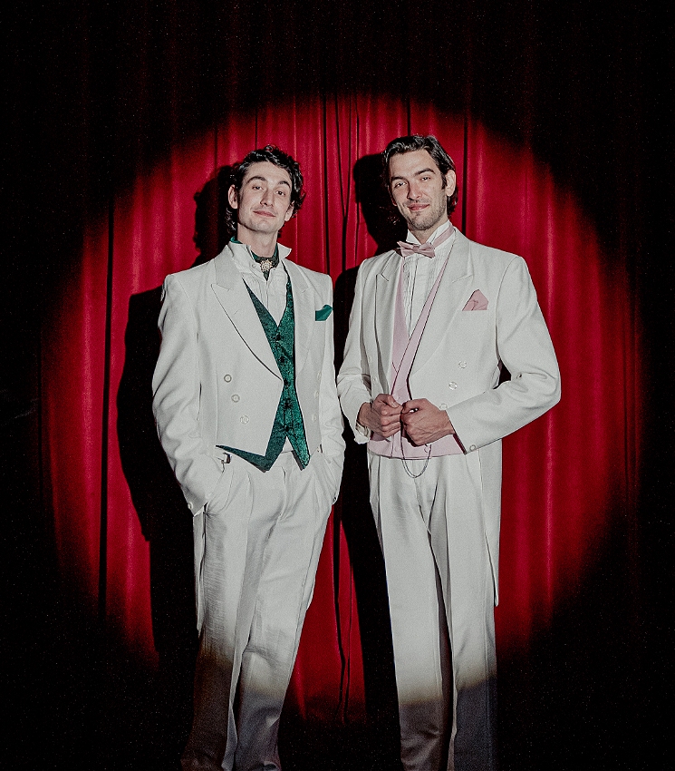 Two brothers stand smiling at the camera, wearing white tailcoat suits in front of a spotlight-lit red curtain. One brother wears a green waistcoat and bowtie, while the other's is pink.